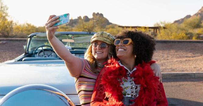 Dos mujeres en un descapotable haciéndose un selfie con el móvil en un paisaje desértico, ilustrando la captura de recuerdos de viajes y la importancia de saber qué es la nube para poder guardar tus fotos gratis sin perder espacio de almacenamiento.