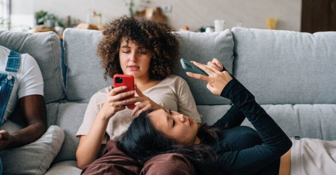 Una mujer sentada en un sofá junto a su amiga recostada en su regazo, ambas utilizando concentradas sus teléfonos móviles, ilustrando el momento de descubrir y compartir los mejores trucos de WhatsApp para hacer la vida más fácil.