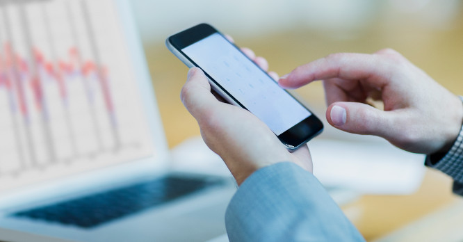 Persona usando su móvil frente a un ordenador con gráficos financieros, representando el funcionamiento digital y el seguimiento de las criptomonedas.