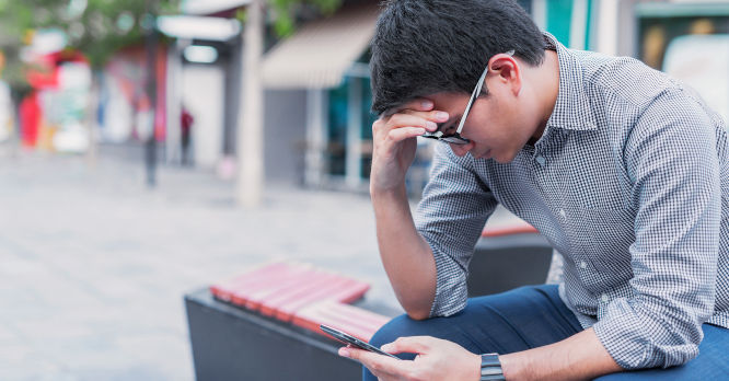 Hombre joven sentado en un banco público con expresión de agobio y cansancio, sujetando su frente con la mano mientras mira su smartphone, ilustrando los efectos de la fatiga del ping y la saturación mental por notificaciones constantes.