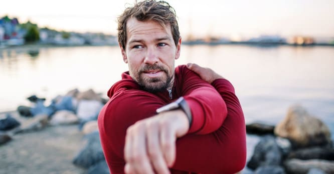 Hombre de mediana edad con barba haciendo estiramientos al aire libre en un entorno costero, llevando un reloj inteligente en su muñeca, que ilustra las funciones de deporte y salud de un smartwatch para monitorear tu actividad.