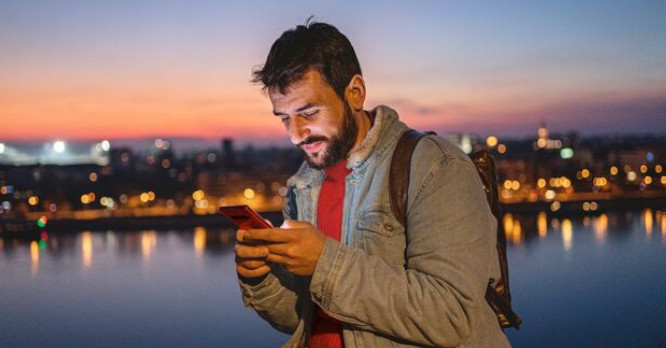 Hombre joven con barba mirando la pantalla iluminada de su móvil al atardecer frente a la ciudad, representando el uso inteligente y los trucos de expertos en WhatsApp para ahorrar tiempo en la comunicación diaria.