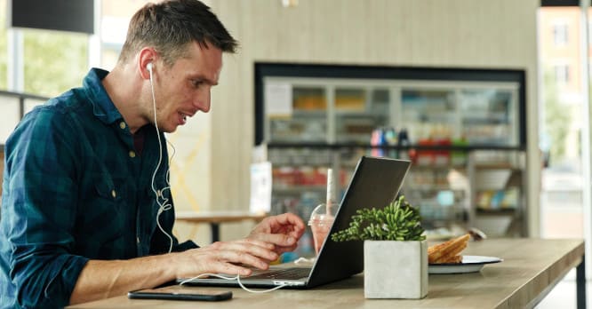 Hombre joven sentado frente a su ordenador portátil con auriculares puestos y un smartphone sobre la mesa, representando la necesidad de aplicar consejos para aumentar tu seguridad digital y proteger tus datos al conectarte desde lugares públicos.
