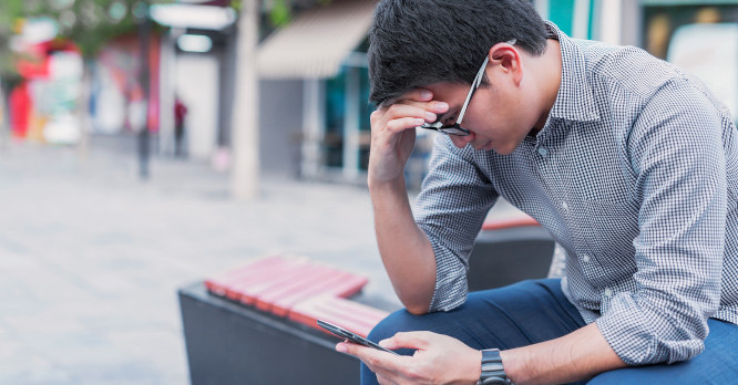 Hombre joven sentado en un banco mirando su teléfono móvil con expresión de preocupación y estrés, ilustrando el impacto negativo de las fake news y lo importante que es aprender a no caer en la desinformación.