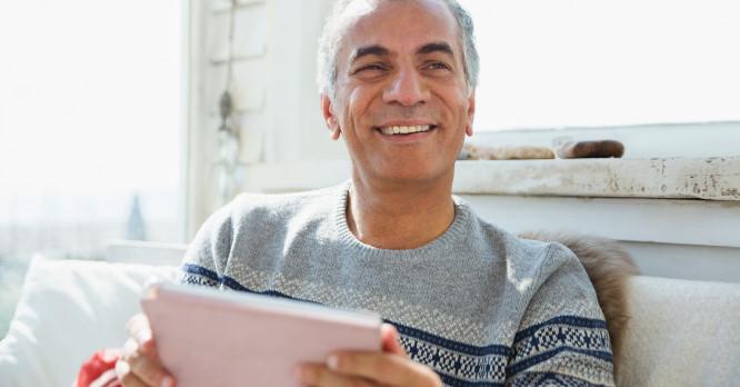 Hombre mayor sonriendo relajadamente en un salón de casa muy luminoso mientras sostiene una tablet, ilustrando la sencillez y las ventajas de controlar un hogar inteligente y automatizar la rutina diaria.