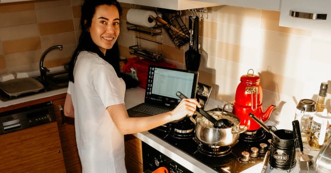 Mujer sonriente cocinando en su cocina junto a un ordenador portátil abierto con una tabla de datos, ilustrando la planificación de comidas en casa y la utilidad de conocer las mejores apps para controlar tu dieta y analizar alimentos de forma sencilla.