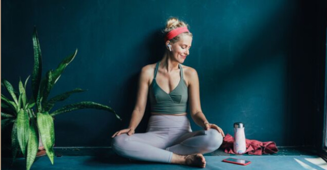 Mujer joven sonriendo en una pose de yoga sobre una esterilla, con auriculares inalámbricos y con su teléfono móvil, ilustrando una rutina de entrenamiento y meditación guiada con las mejores apps para cuidar tu salud desde tu móvil.