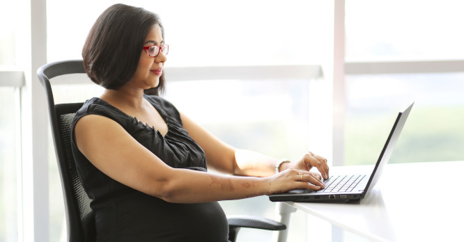 Mujer embarazada de perfil con gafas rojas trabajando frente a un ordenador portátil en una mesa iluminada por el sol, ilustrando cómo aplicar los consejos de expertos para proteger tus ojos de la luz de las pantallas y evitar la fatiga visual.