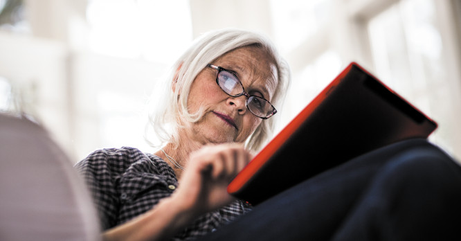 Primer plano de una mujer mayor con gafas mirando concentrada la pantalla de su tableta con funda roja, representando el aprendizaje digital y la importancia de ayudar a nuestros padres a protegerse de las estafas digitales e internet.