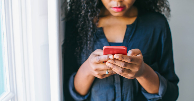 Una mujer de pelo rizado sosteniendo y escribiendo en un teléfono móvil con funda de color rojo cerca de una ventana, ilustrando la consulta de aplicaciones y la importancia de seguir una guía rápida para liberar espacio en tu móvil Android e iOS.