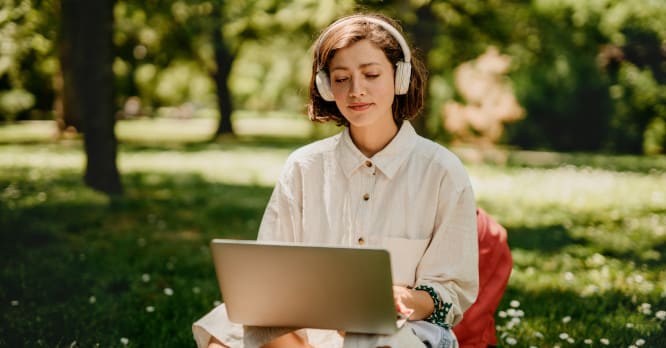 Mujer joven sentada en el césped de un parque navegando en su portátil con unos auriculares de diadema blancos, ilustrando cómo disfrutar de la tecnología manteniendo hábitos seguros de volumen y tiempo para proteger la salud auditiva.