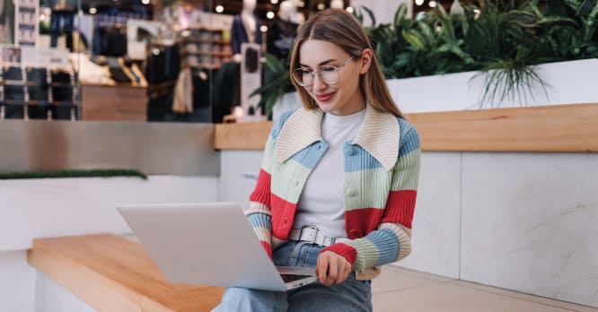 Mujer joven conectada a su ordenador portátil desde un espacio público comercial, ilustrando los hábitos de navegación diaria y la importancia de saber qué es la huella digital y cómo protegerla.