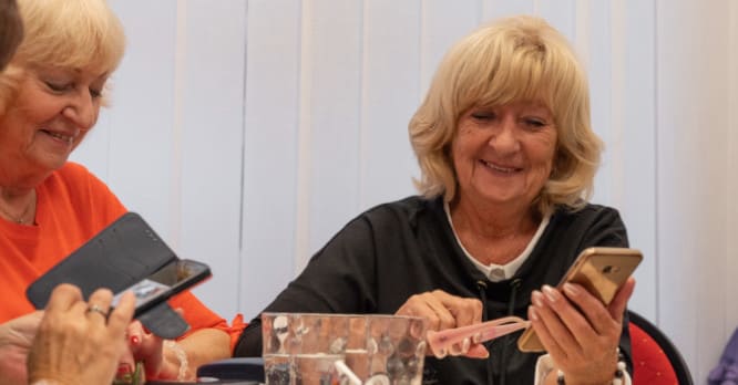 Dos mujeres mayores sonriendo mientras aprenden a usar sus teléfonos móviles, ilustrando la importancia de la educación tecnológica para superar la brecha digital y ayudar a la tercera edad.