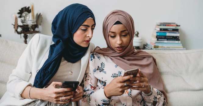 Dos mujeres jóvenes sentadas en un sofá mirando con atención la pantalla de un móvil, ilustrando el proceso de revisar la privacidad, hacer copias de seguridad o transferir datos correctamente antes de reciclar o vender tu móvil.