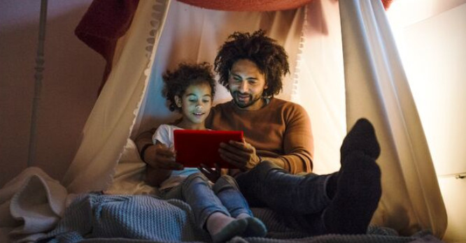 Un padre y su hija pequeña sonriendo juntos dentro de una cabaña de mantas iluminados por el brillo de una pantalla de tablet, representando el acompañamiento activo y cómo proteger a los menores cuando navegan por internet de manera segura.