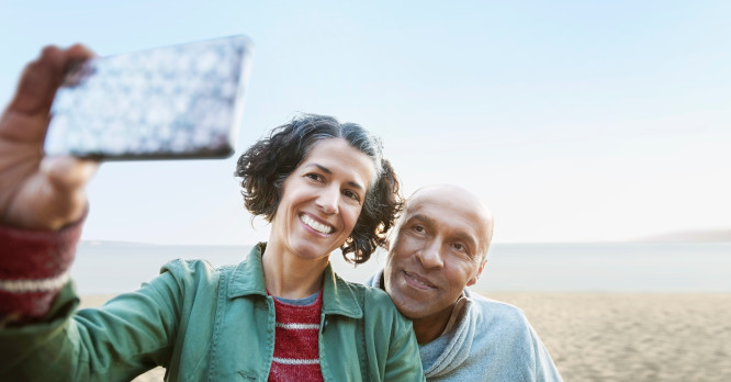Pareja de mediana edad sonriendo mientras se hacen un selfie con el móvil en la playa al atardecer, representando los momentos de descanso y la necesidad de aplicar pautas de ciberseguridad en vacaciones para proteger nuestra información al viajar.