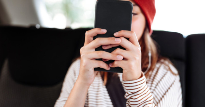 Joven en un coche mirando con mucha atención la pantalla de su teléfono móvil con auriculares puestos, ilustrando el consumo intensivo de recursos y la importancia de seguir buenos hábitos para que la batería del teléfono dure más tiempo.
