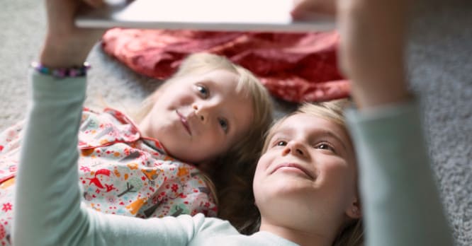 Dos niñas pequeñas miran una tableta que sostienen sobre ellas, una escena que ilustra la importancia de acompañar a los menores y hablar sobre el uso responsable y seguro de los dispositivos digitales.